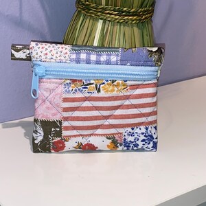 May include: A small quilted zippered pouch with a patchwork design featuring various floral and geometric patterns in shades of pink, blue, green, and white.