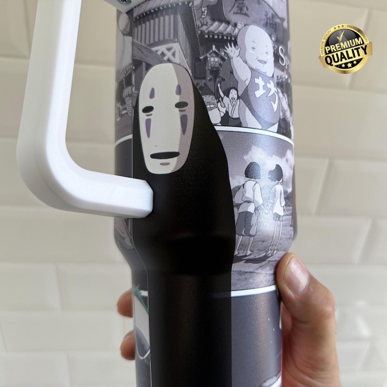 May include: A black and white tumbler featuring imagery from the animated film Spirited Away. The tumbler has a white handle and a black base. The design includes the character No-Face and other scenes from the movie. A gold "Premium Quality" sticker is visible.