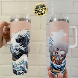 May include: A large insulated travel mug with a white handle and straw. The mug features a Japanese art-inspired design of a wave in blue and white. The top of the mug has a pink and white gradient. The mug is designed for cold drinks.