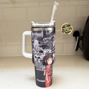 May include: A black and white tumbler featuring panels from the animated film "Spirited Away." The tumbler has a white handle and a clear lid with a straw. A "Premium Quality" sticker is visible on the right.