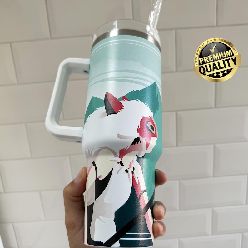 May include: A large, insulated tumbler with a white handle and a straw. The tumbler features a design of a character with white fur, a red mask, and a mountain landscape in shades of teal and green. A gold "Premium Quality" sticker is visible.