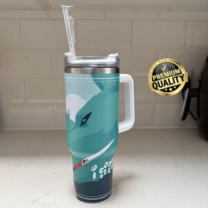 May include: A teal and silver insulated travel mug with a white handle and lid. The mug features a graphic of a wolf-like creature and a white straw. A gold "Premium Quality" sticker is on the right.