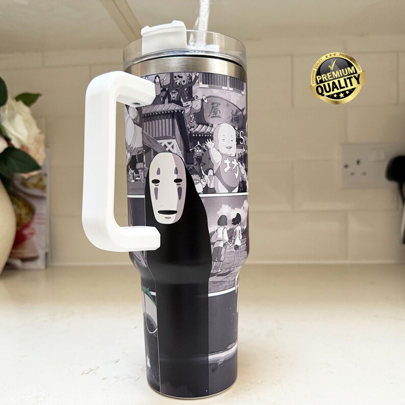 May include: A stainless steel tumbler with a white handle and straw. The tumbler features a black and white graphic of characters from the animated film "Spirited Away". A "Premium Quality" sticker is visible.