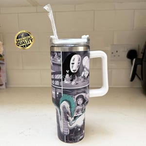May include: A stainless steel tumbler with a white handle and lid, featuring a black and white manga-style illustration. The design includes characters and scenes from an animated film. A clear straw extends from the lid.