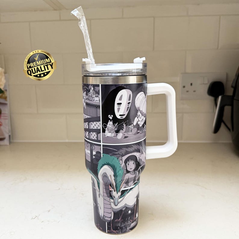May include: A stainless steel tumbler with a white handle and lid, featuring a black and white manga-style illustration. The design includes characters and scenes from an animated film. A clear straw extends from the lid.