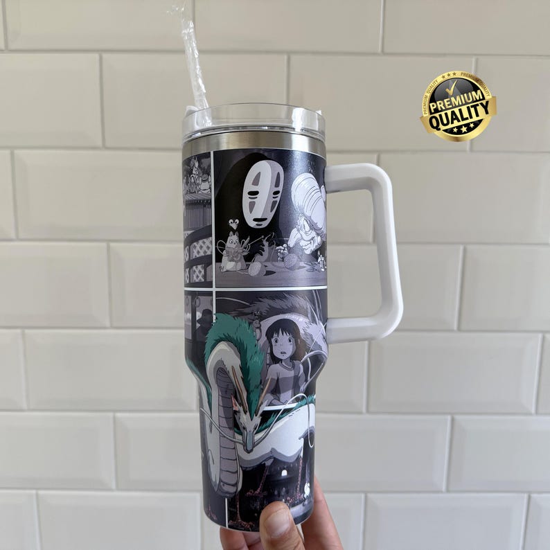 May include: A tall, stainless steel tumbler with a white handle and a clear lid. The tumbler is decorated with a black and white manga-style graphic featuring characters and scenes. A clear straw is inserted into the lid.