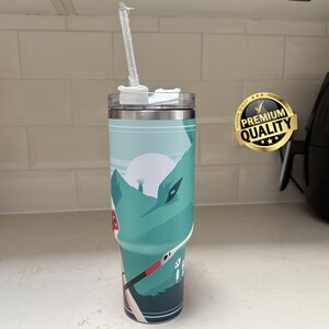 May include: A stainless steel tumbler with a teal and white mountain scene design. The tumbler has a clear lid and a clear straw. A gold "Premium Quality" sticker is on the right side.