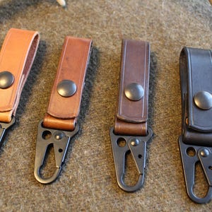 Puede incluir: Cuatro llaveros de cuero en tonos beige, marrón y negro. Cada llavero tiene un cierre de metal y un cierre a presión. Los llaveros se muestran sobre una superficie marrón texturizada.