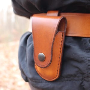Puede incluir: Una funda de cuero marrón sujeta a un cinturón de cuero marrón. La funda tiene forma curva y cierre de botón a presión. El cinturón es ancho y de diseño sencillo. El fondo está borroso, lo que sugiere un entorno al aire libre.