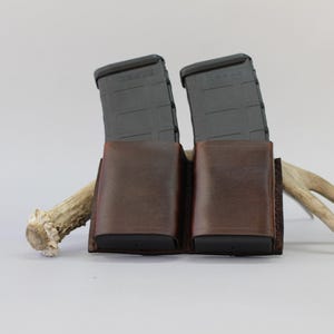 May include: A dark brown leather double magazine pouch holding two black polymer rifle magazines. The pouch is positioned in front of a light-colored deer antler, featuring visible stitching along the edges and a smooth, polished finish on the leather.