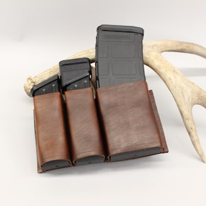 以下が含まれることがあります： 異なる高さの黒いポリマー製銃器用マガジン3本を収納した茶色のレザー製トリプルマガジンポーチ。ポーチはステッチが施されたエッジと構造化されたデザインが特徴で、各マガジンを個別に収納できるコンパートメントが並んでいます。