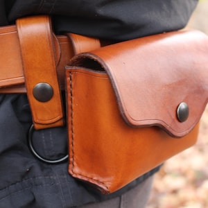 Puede incluir: Un cinturón de cuero marrón con una bolsa a juego adjunta. La bolsa tiene una solapa curva y un cierre a presión. El cinturón y la bolsa están hechos de cuero rico y cálido. La bolsa mide aproximadamente 15 cm de ancho.