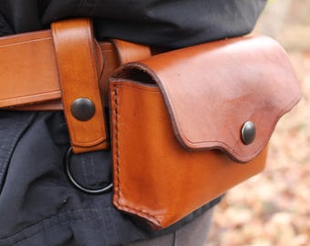 Puede incluir: Un cinturón de cuero color canela con una bolsa de cuero a juego en el lateral. La bolsa tiene una solapa curva asegurada con un botón de presión de metal oscuro. Una anilla en D de metal oscuro está unida a la trabilla, y el cinturón se cierra.