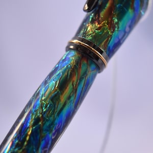 K&ouml;nnte beinhalten: Nahaufnahme eines F&uuml;llfederhalters mit einem lebendigen, schillernden Schaft. Der Stiftk&ouml;rper weist ein wirbelndes Muster aus Blau, Gr&uuml;n und Orange auf, das durch ein schwarzes Band mit goldener Zierleiste akzentuiert wird. Das Design des Stifts ist elegant und auff&auml;llig.