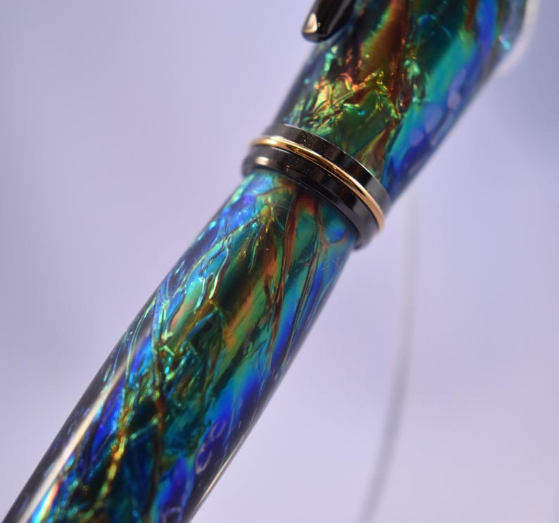 K&ouml;nnte beinhalten: Nahaufnahme eines F&uuml;llfederhalters mit einem lebendigen, schillernden Schaft. Der Stiftk&ouml;rper weist ein wirbelndes Muster aus Blau, Gr&uuml;n und Orange auf, das durch ein schwarzes Band mit goldener Zierleiste akzentuiert wird. Das Design des Stifts ist elegant und auff&auml;llig.