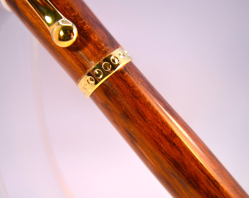 K&ouml;nnte beinhalten: Nahaufnahme eines Holzstiftes mit einem goldenen Band und einer goldenen Kappe. Der Stift hat eine dunkelbraune Holzmaserung.