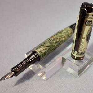 May include: A distinctive fountain pen with a money-themed design. The pen's barrel is patterned to resemble U.S. currency, and the cap incorporates a section of a hundred-dollar bill. It has a dark metal clip and nib, displayed on a clear acrylic stand.