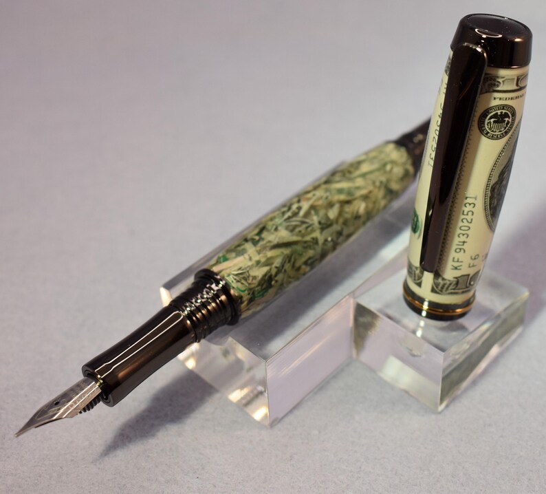 May include: A distinctive fountain pen with a money-themed design. The pen's barrel is patterned to resemble U.S. currency, and the cap incorporates a section of a hundred-dollar bill. It has a dark metal clip and nib, displayed on a clear acrylic stand.