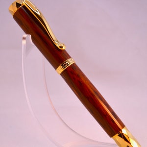 K&ouml;nnte beinhalten: Ein brauner und goldener Stift mit Holzmaserung und einem goldenen Band mit einem sich wiederholenden Kreisdesign. Der Stift befindet sich auf einem durchsichtigen Acrylst&auml;nder.