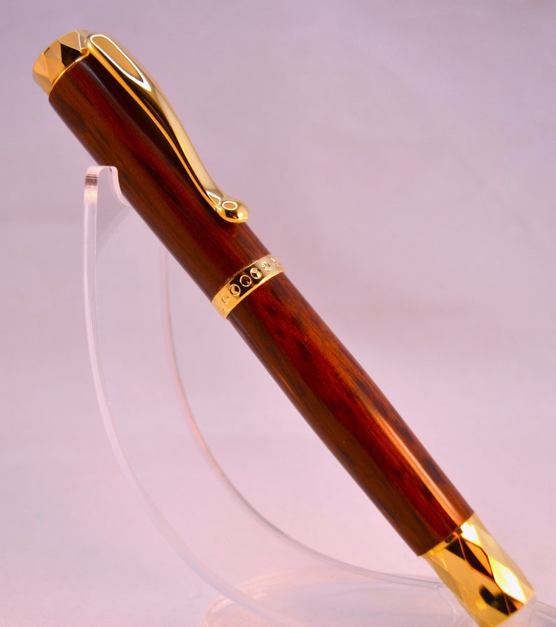 K&ouml;nnte beinhalten: Ein brauner und goldener Stift mit Holzmaserung und einem goldenen Band mit einem sich wiederholenden Kreisdesign. Der Stift befindet sich auf einem durchsichtigen Acrylst&auml;nder.