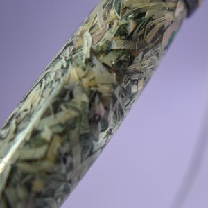 May include: A pen with a clear barrel filled with shredded banknotes. The pen has a dark cap and a silver-coloured tip. The background is a soft purple.