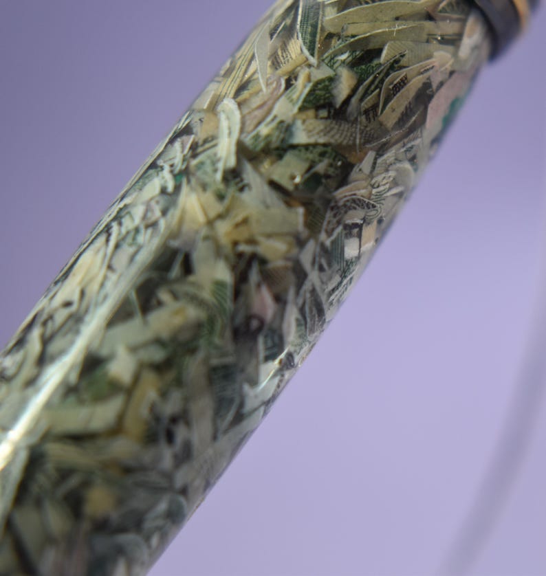 May include: A pen with a clear barrel filled with shredded banknotes. The pen has a dark cap and a silver-coloured tip. The background is a soft purple.
