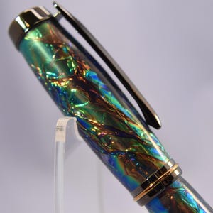 K&ouml;nnte beinhalten: Nahaufnahme eines Stifts mit einem schimmernden, irisierenden Schaft in Gr&uuml;n-, Blau- und Goldt&ouml;nen. Der Stift hat einen silbernen Clip und einen goldenen Ring in der N&auml;he der Spitze. Der Stift wird auf einem transparenten Acrylst&auml;nder pr&auml;sentiert.