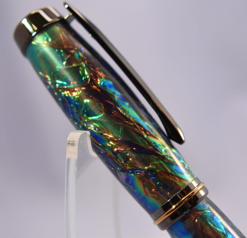 K&ouml;nnte beinhalten: Nahaufnahme eines Stifts mit einem schimmernden, irisierenden Schaft in Gr&uuml;n-, Blau- und Goldt&ouml;nen. Der Stift hat einen silbernen Clip und einen goldenen Ring in der N&auml;he der Spitze. Der Stift wird auf einem transparenten Acrylst&auml;nder pr&auml;sentiert.