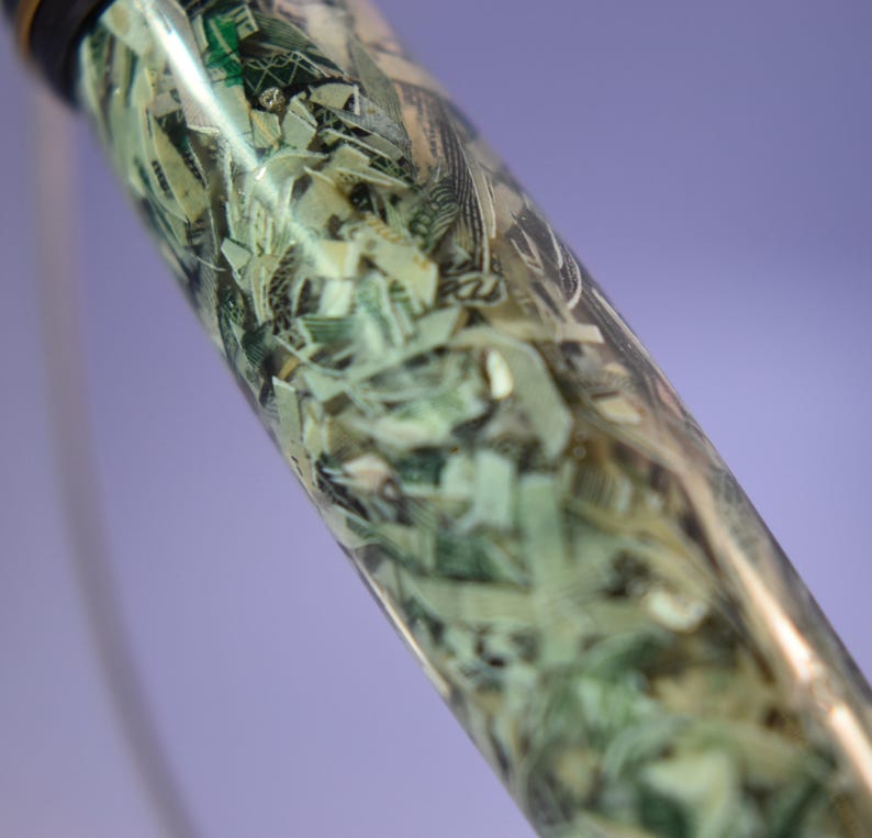 May include: A clear, cylindrical object filled with shredded U.S. dollar bills. The money is visible through the transparent material, creating a unique texture. The top of the object is black.