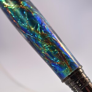 K&ouml;nnte beinhalten: Nahaufnahme eines Stifts mit schimmerndem, irisierendem Schaft. Der Schaft weist ein strukturiertes, mehrfarbiges Design mit Blau-, Gr&uuml;n- und Goldt&ouml;nen auf. Die Spitze und die Akzente des Stifts sind dunkel und metallisch.