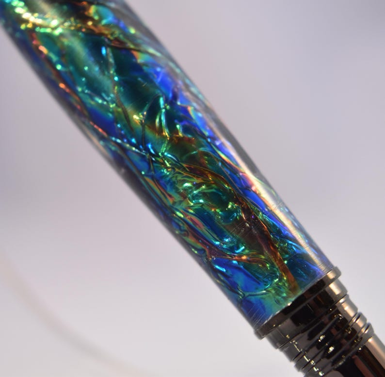 K&ouml;nnte beinhalten: Nahaufnahme eines Stifts mit schimmerndem, irisierendem Schaft. Der Schaft weist ein strukturiertes, mehrfarbiges Design mit Blau-, Gr&uuml;n- und Goldt&ouml;nen auf. Die Spitze und die Akzente des Stifts sind dunkel und metallisch.