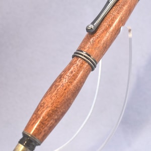 May include: A wooden pen with a reddish-brown hue, complemented by silver-coloured metal accents and a clip. The pen's design is highlighted by a clear acrylic stand. The pen's overall length is approximately 14 cm.
