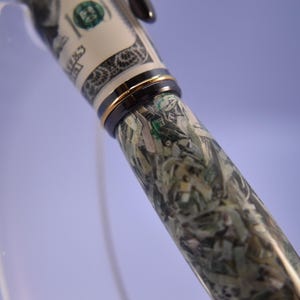 May include: A pen with a design resembling a hundred-dollar bill. The pen has a black and gold band near the tip and a black top. The pen's barrel is covered in a pattern of green, white, and black, mimicking shredded currency. The pen is a writing instrument.