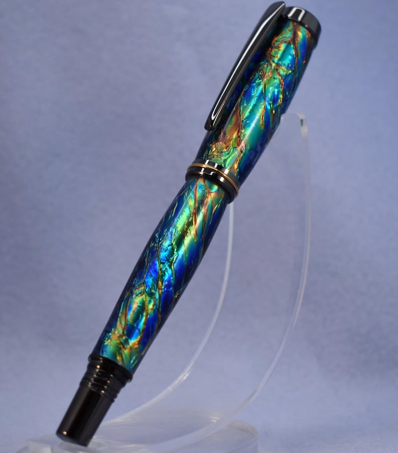 K&ouml;nnte beinhalten: Ein lebendiger F&uuml;llfederhalter mit einem Wirbelmuster aus Blau, Gr&uuml;n und Gold. Der Stift hat einen schwarzen Clip und Akzente sowie ein goldfarbenes Band. Der Stift wird auf einem transparenten St&auml;nder ausgestellt.