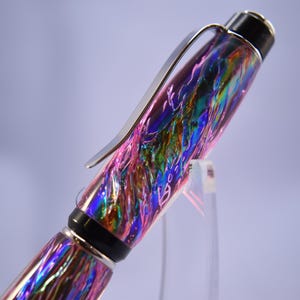 May include: A colorful pen with a swirling, iridescent design in shades of pink, blue, green, and purple. The pen has a silver clip and black accents. The pen is displayed on a clear stand.