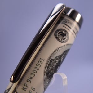 May include: A close-up of a pen with a silver clip and a barrel designed to look like a US $100 bill. The pen has the serial number KF 94302531 printed on it. The pen is displayed on a clear stand against a purple background.