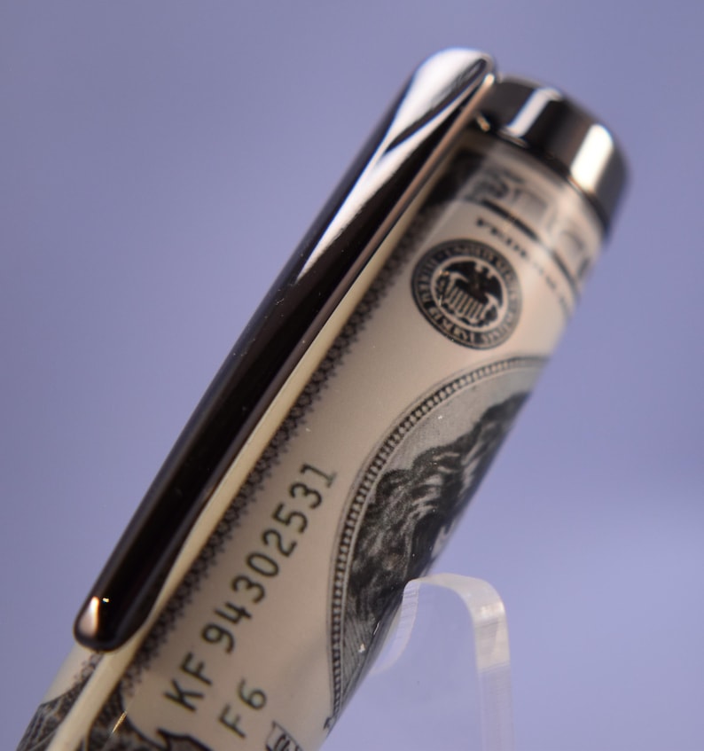 May include: A close-up of a pen with a silver clip and a barrel designed to look like a US $100 bill. The pen has the serial number KF 94302531 printed on it. The pen is displayed on a clear stand against a purple background.