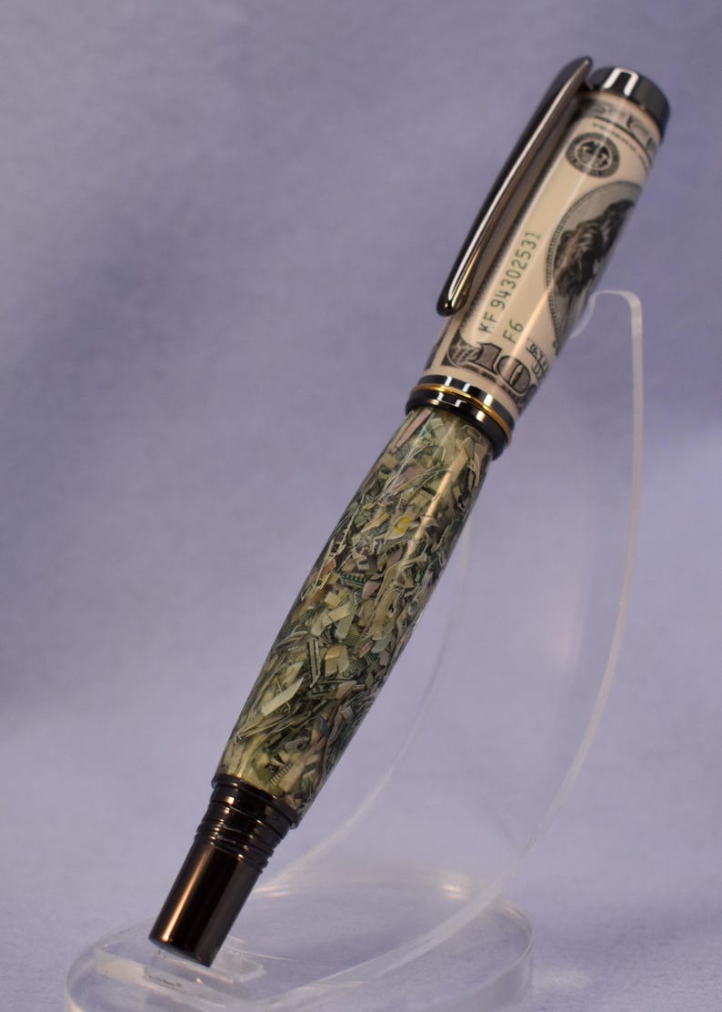 May include: A distinctive pen with a design incorporating elements of a US $100 bill. The pen has a black clip and tip, with a decorative band. The barrel is a mosaic of green and tan fragments, creating a textured appearance.