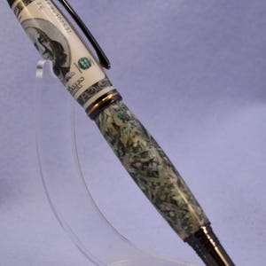 May include: A distinctive pen with a design inspired by a US $100 bill. The pen has a black clip and tip, with a barrel that appears to be made of green and white material. The pen is displayed on a clear stand.