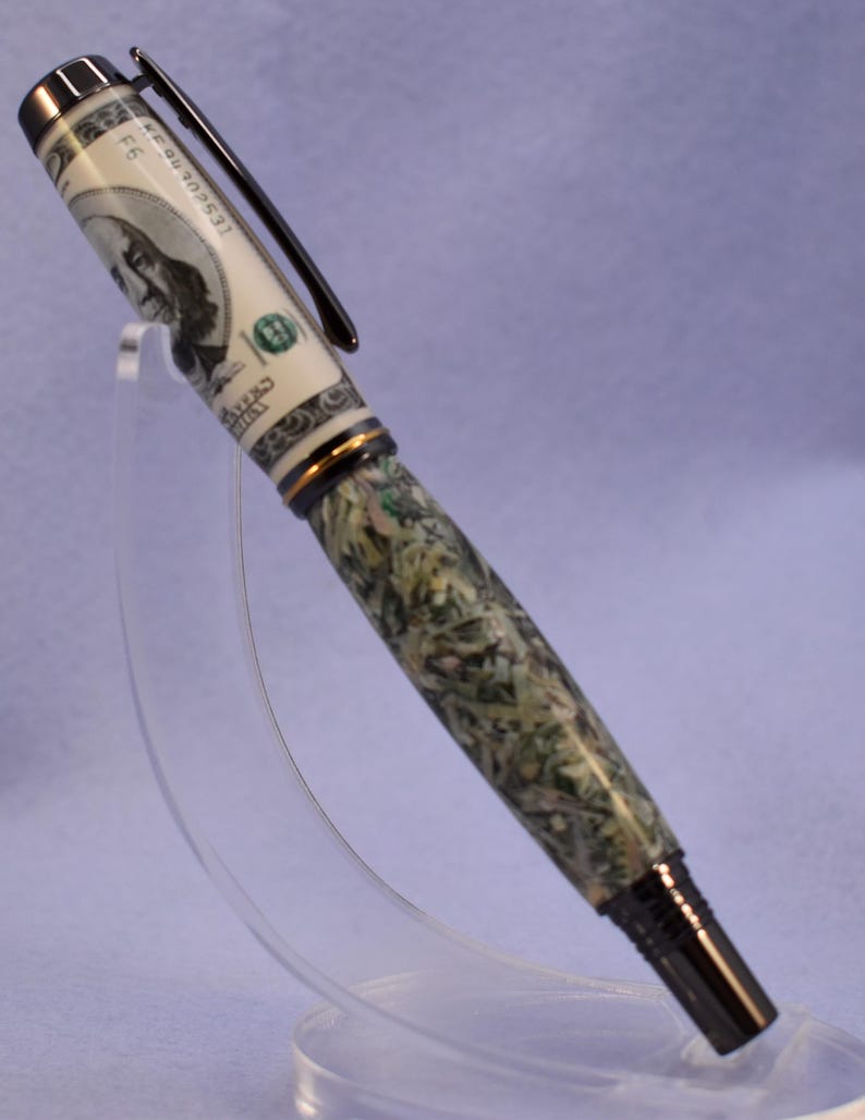 May include: A distinctive pen with a design inspired by a US $100 bill. The pen has a black clip and tip, with a barrel that appears to be made of green and white material. The pen is displayed on a clear stand.