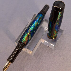 K&ouml;nnte beinhalten: Ein F&uuml;llfederhalter mit schwarzem Korpus und Kappe. Der Stift hat ein farbenfrohes, schillerndes Design mit Blau-, Gr&uuml;n- und Goldt&ouml;nen. Die Feder ist goldfarben. Die Kappe ist abgenommen und zeigt das Design des Stifts.