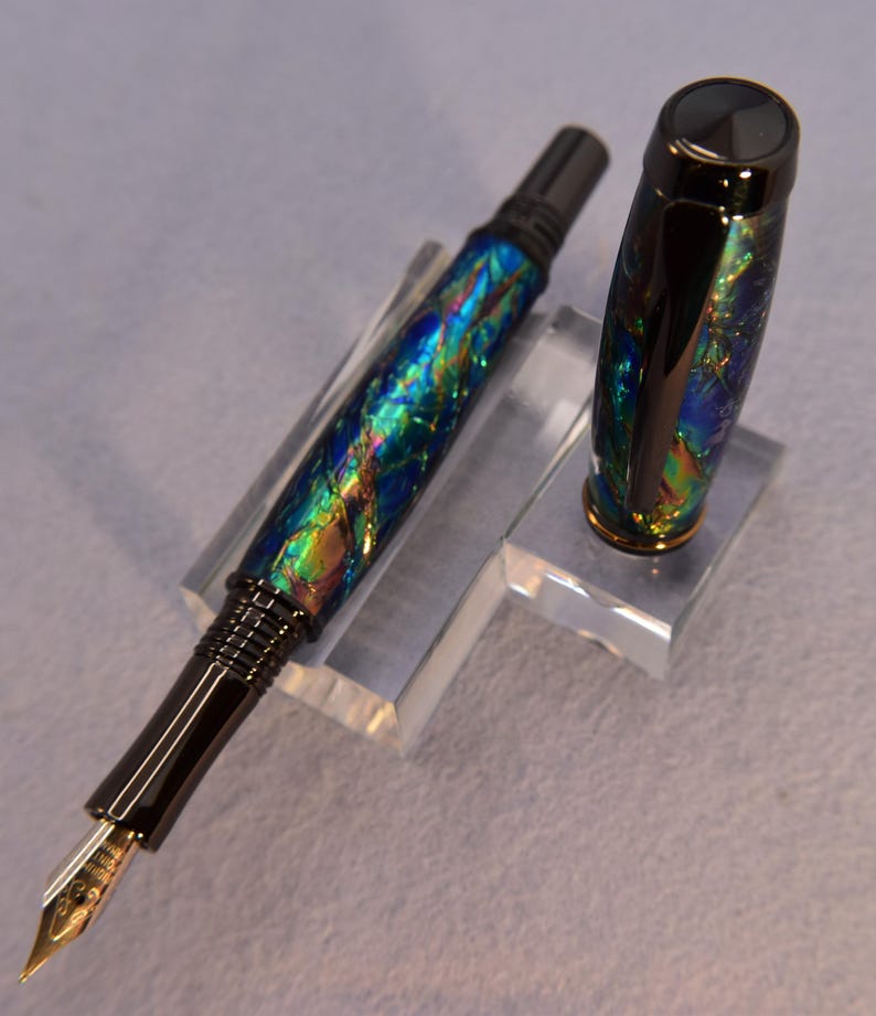 K&ouml;nnte beinhalten: Ein F&uuml;llfederhalter mit schwarzem Korpus und Kappe. Der Stift hat ein farbenfrohes, schillerndes Design mit Blau-, Gr&uuml;n- und Goldt&ouml;nen. Die Feder ist goldfarben. Die Kappe ist abgenommen und zeigt das Design des Stifts.