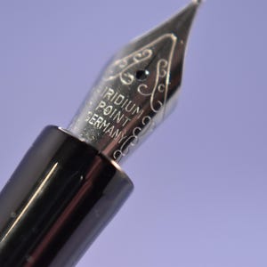 May include: Close-up of a fountain pen nib with the text "IRIDIUM POINT GERMANY". The nib is silver-coloured with decorative scrollwork. The pen's body is black and glossy, set against a soft purple background.