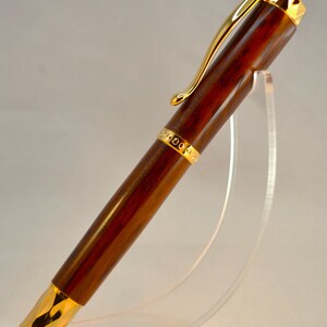 K&ouml;nnte beinhalten: Ein brauner Holzstift mit goldenen Akzenten und einer goldenen Klammer. Der Stift steht auf einem durchsichtigen Acrylst&auml;nder.