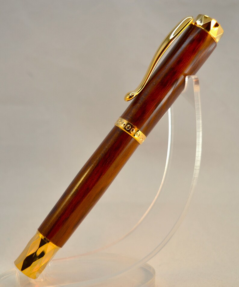 K&ouml;nnte beinhalten: Ein brauner Holzstift mit goldenen Akzenten und einer goldenen Klammer. Der Stift steht auf einem durchsichtigen Acrylst&auml;nder.