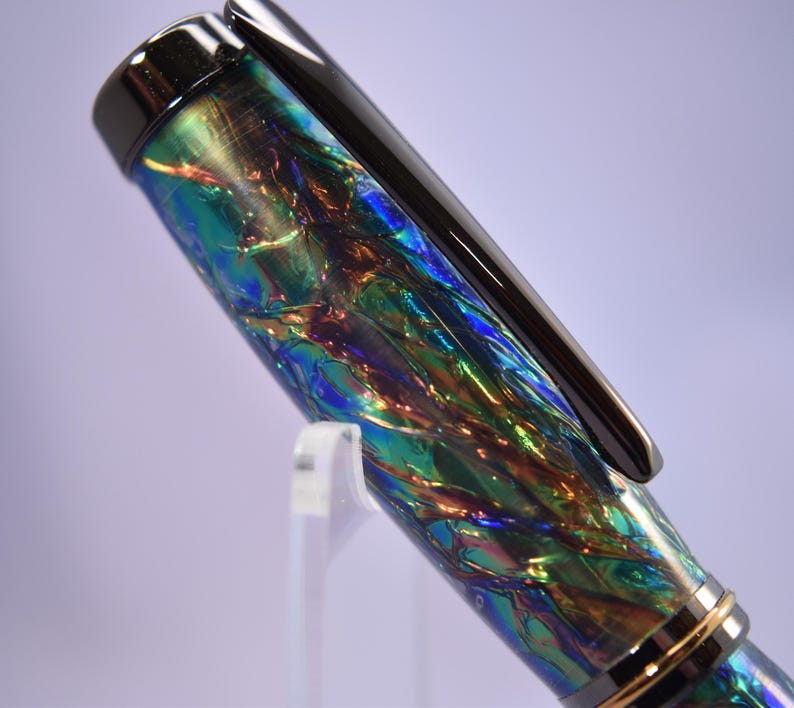 K&ouml;nnte beinhalten: Nahaufnahme eines F&uuml;llfederhalters mit schimmerndem, irisierendem Schaft. Der Stift weist eine Mischung aus blauen, gr&uuml;nen und goldenen Farben auf. Ein schwarzer Clip und goldene Akzente zieren den Stift, der leicht nach links geneigt ist.