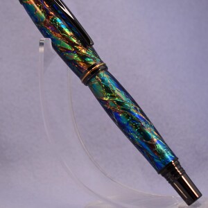 K&ouml;nnte beinhalten: Ein F&uuml;llfederhalter mit einem lebendigen, schillernden Finish in Blau-, Gr&uuml;n- und Goldt&ouml;nen. Der Stift hat einen schwarzen Clip und eine Spitze sowie ein goldenes Band in der Mitte. Er wird auf einem transparenten St&auml;nder pr&auml;sentiert.