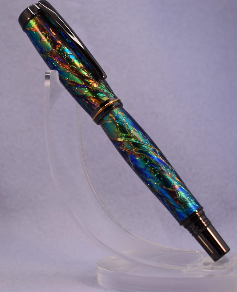 K&ouml;nnte beinhalten: Ein F&uuml;llfederhalter mit einem lebendigen, schillernden Finish in Blau-, Gr&uuml;n- und Goldt&ouml;nen. Der Stift hat einen schwarzen Clip und eine Spitze sowie ein goldenes Band in der Mitte. Er wird auf einem transparenten St&auml;nder pr&auml;sentiert.