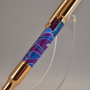 May include: A gold and purple pen with a swirling pattern. The pen is made from a bullet casing and has a gold clip.