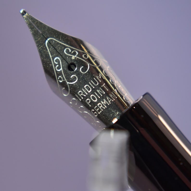 K&ouml;nnte beinhalten: Nahaufnahme einer silbernen F&uuml;llfederhalters mit der Gravur "IRIDIUM POINT GERMANY". Die Feder hat dekorative Schn&ouml;rkel und ein kleines Loch. Der schwarze Schaft des Stifts ist im Hintergrund zu sehen.